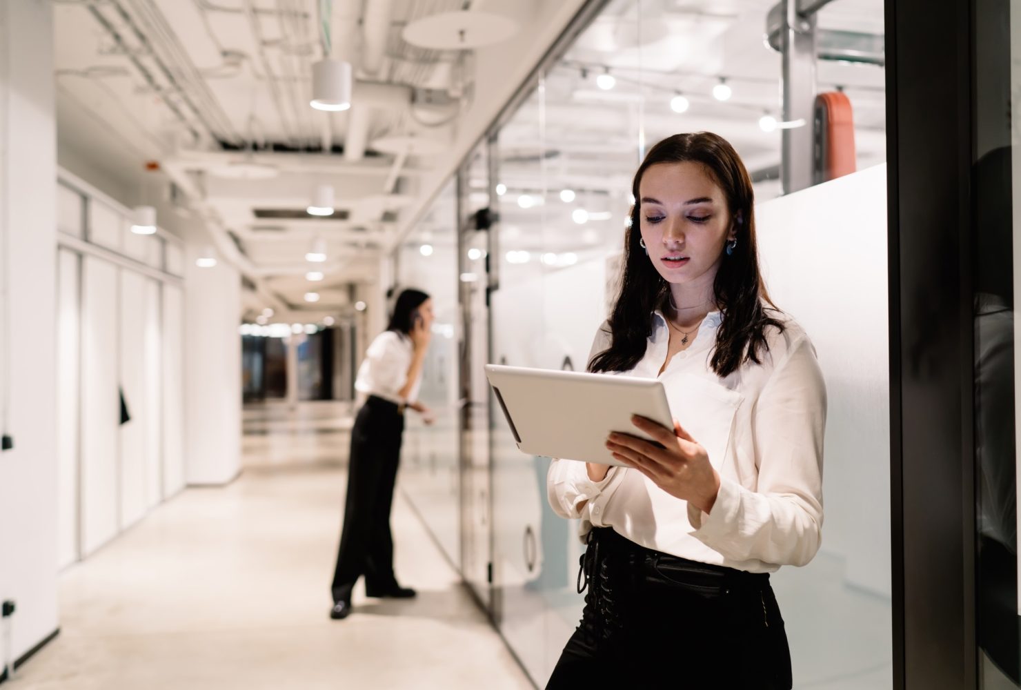 Cheerful young businesswoman standing in office corridor with tablet and looking at screen
